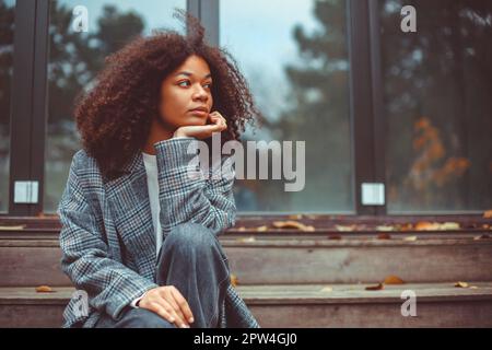 Portrait d'automne extérieur de la jeune femme afro-américaine attentionnés regardant de côté assis sur les escaliers de maison en bois vieux, pensive fille curly passant du temps Banque D'Images