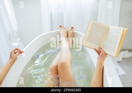 Vue arrière de la jeune femme brunette lisant le livre et buvant du vin blanc dans le bain avec de la mousse et des bougies Banque D'Images