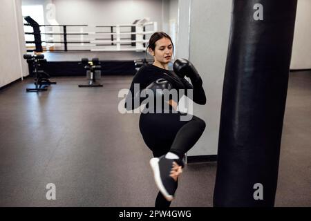 Une femme de boxe latine forte saute de coup de pied à un énorme sac de poinçonnage à la salle de fitness. Entraînement de fille athlétique Muay Thai boxe pour le culturisme et la santé Banque D'Images