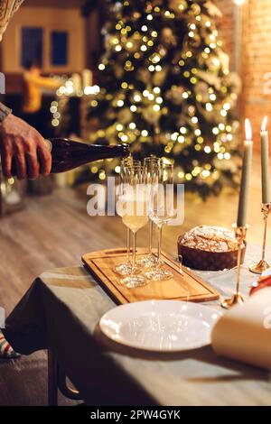Célébration de la Saint-Sylvestre. Homme versant du champagne dans des verres debout sur la table avec un dîner de Noël festif, des bougies et des cadeaux enveloppés contre flou Banque D'Images