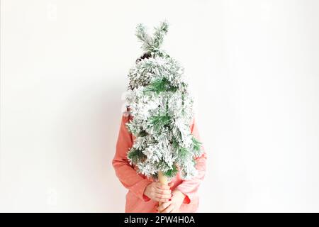 Femme tenant un petit arbre de Noël devant le visage tout en se tenant isolée sur fond blanc de studio. Femelle avec fausse couche de neige artificielle Banque D'Images