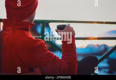 Homme en vêtements d'hiver buvant du thé chaud à base de plantes. Soirée d'hiver depuis un beau balcon à l'heure du suset sur le lac Baikal. Le tourisme d'hiver Banque D'Images