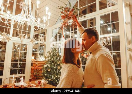 Baiser de minuit. Un jeune couple de familles heureux, amoureux, embrasse et embrasse pendant la fête de la Saint-Sylvestre dans une chambre confortable décorée avec un arbre de Noël Banque D'Images