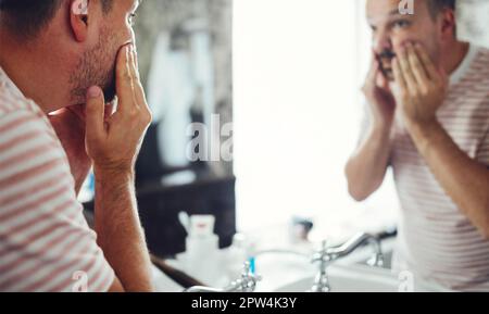 Homme beau millénaire regardant dans le miroir, appliquant de la crème hydratante sur les joues dans la salle de bains, gros plan de la tête. Jeune garçon bien entretenu Banque D'Images