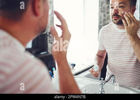 Homme beau millénaire regardant dans le miroir, appliquant hydratant après le rasage de la crème sur les joues dans la salle de bains, coup de tête gros plan. Bien entretenu jeune Banque D'Images