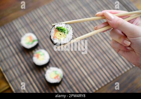 Une main avec des baguettes est titulaire d'un sushi roll sur un tapis de paille bambou serwing arrière-plan. Cuisine asiatique traditionnelle. Banque D'Images
