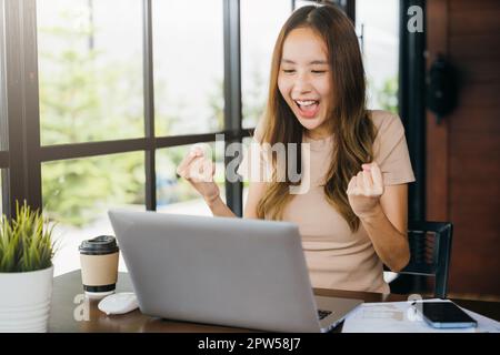 Une jeune femme d'affaires asiatique heureuse et excitée, assise avec un ordinateur portable au comptoir du café, se lève les mains et célèbre le succès, la femme se sentant gagnante réj Banque D'Images