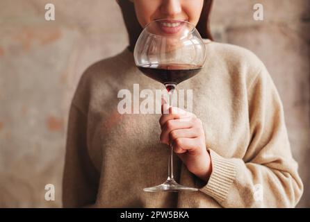 Crop Happy female en chandail sentant le vin rouge dans un grand verre sur fond gris Banque D'Images