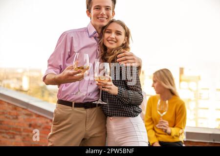 Joyeux jeune couple amoureux s'embrassant sur la terrasse de la maison, tenant des verres de vin blanc, souriant et posant sur l'appareil photo, l'homme et son bien-aimé Banque D'Images