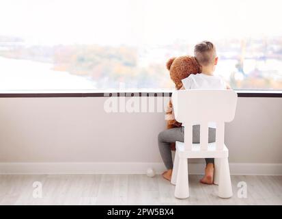 Vue arrière d'un petit garçon avec un ours en peluche assis sur une chaise et regardant par la fenêtre dans le paysage urbain d'automne tout en attendant la maman à la maison.Concept de l'enfance Banque D'Images