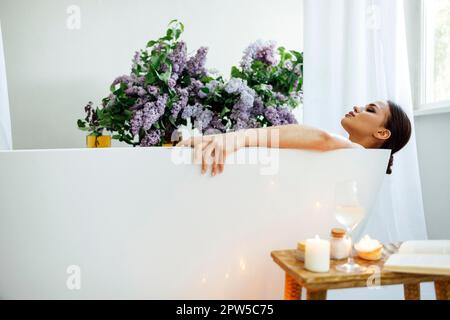 Vue latérale de la jeune femme brune lisant le livre et buvant du vin blanc dans le bain avec de la mousse et des bougies Banque D'Images