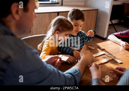 La mère, le père et les enfants jouent aux dominos tout en étant assis dans le salon près de la fenêtre. Famille passant du temps ensemble Banque D'Images