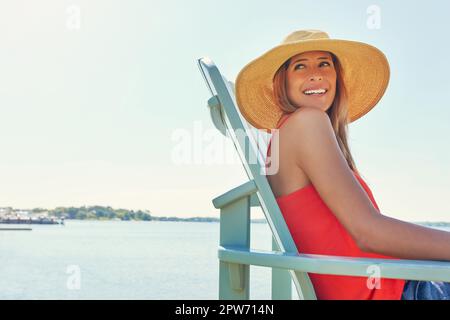 Elle aime vivre près de l'eau. une jeune femme joyeuse portant un chapeau tout en étant assise sur une chaise près d'un lac à l'extérieur au soleil. Banque D'Images