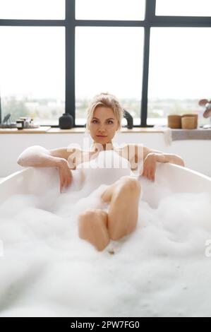 Portrait d'une femme détendue profitant de la journée spa tout en étant allongé dans la mousse de bain à la salle de bains à la maison avec de la mousse de savon, des bulles et regardant l'appareil photo. Beauté et corps ca Banque D'Images