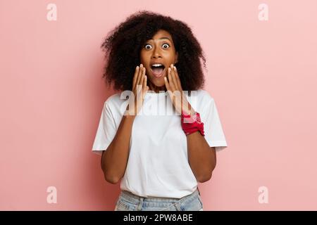 Femme dans une émotion surprise, elle se tient vêtue d'un t-shirt blanc sur fond rose. Photo de haute qualité Banque D'Images
