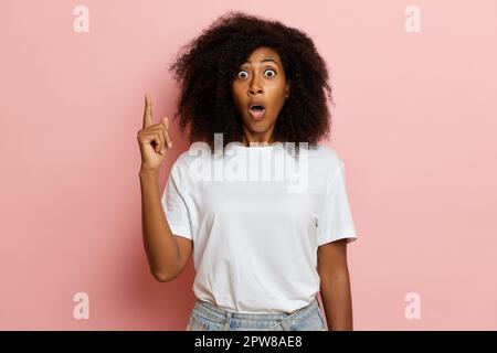 Une femme à la peau foncée montre le doigt vers le haut, elle a ouvert sa bouche dans une surprise et a ouvert ses yeux largement isolés sur le mur rose. Banque D'Images