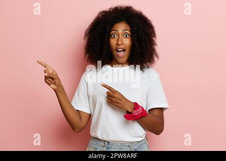 Jolie femme à la peau foncée avec une expression faciale heureuse pointant sur un côté sur fond rose, espace de copie. Banque D'Images