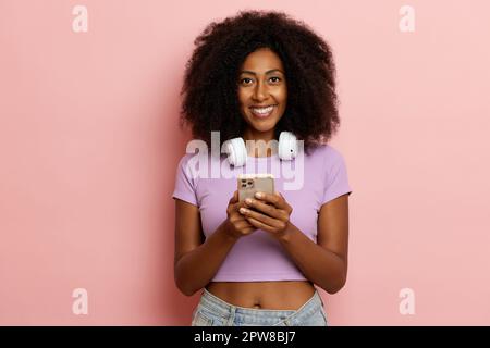 Jolie fille ethnique positive avec des cheveux molletonnés, sourit édroitement et regarde la caméra, utilise le téléphone pour les messages, habillé dans un tissu décontracté, pose ov Banque D'Images