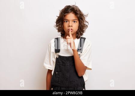 Plan horizontal de mystérieux jeune gars avec des cheveux bouclés fait le geste de silence avec l'expression étourdisse, garde le doigt sur les lèvres, partage secret, porte ca Banque D'Images