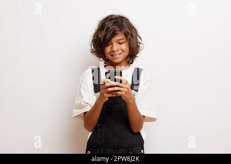 Prise de vue en intérieur d'un jeune garçon positif aux cheveux bouclés, utilise un téléphone portable pour discuter en messagerie, sourires joyeux, porte un tissu décontracté, isolé sur une wa blanche Banque D'Images