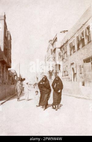 Égypte. Vers.1918. Deux femmes marchant le long d'une rue accompagnées d'un enfant en bas âge. Les femmes portent al-habara, composé d'une jupe pleine longueur, d'une coiffe de tête, et al-burqu», une couverture de visage. Une femme porte également Arūsat al burqa, ornementation portée sur le voile de visage. Deux hommes marchent dans la direction opposée, dont l'un regarde en arrière. Banque D'Images