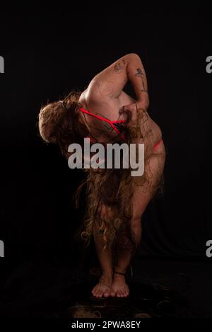 Jeune homme, acteur, portant bikini dans performance gorilla femme avec les cheveux partout dans son corps et expression de la douleur. isolé sur fond sombre. Banque D'Images