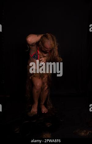 Jeune homme, acteur, portant bikini dans performance gorilla femme avec les cheveux partout dans son corps et expression de la douleur. isolé sur fond sombre. Banque D'Images