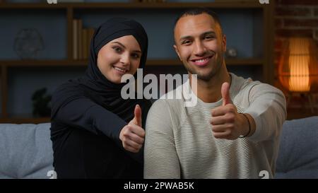 Couple souriant famille africain américain et arabian musulmane femme diverse femme mari assis sur le canapé ensemble montrant les pouces comme positif Banque D'Images