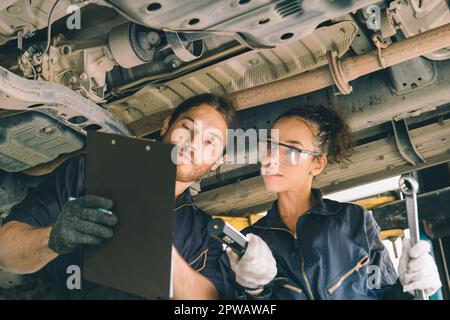 L'homme et la femme de l'équipe de service mécanicien travaillent ensemble sous un palan de voiture liste de vérification de l'entretien de la suspension dans le garage auto wokshop Banque D'Images