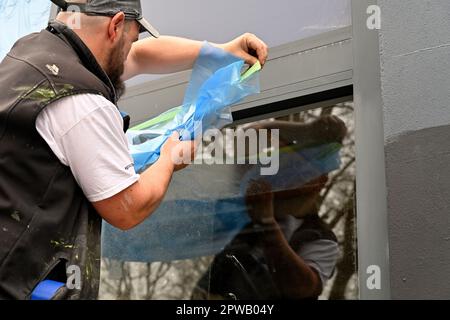 Workman pose une feuille de masquage de protection en plastique sur la fenêtre en verre de l'atelier en vue de la peinture par pulvérisation Banque D'Images