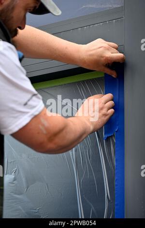 Workman tapissant la feuille de masquage de protection en plastique sur la fenêtre en verre de l'atelier en vue de la peinture par pulvérisation Banque D'Images