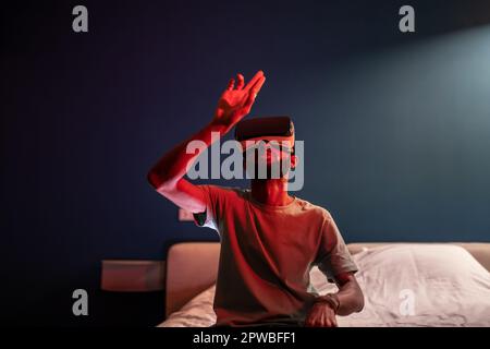 Jeune homme afro-américain assis sur un lit sous une lumière néon rouge à l'aide d'un casque VR métaverse futuriste Banque D'Images
