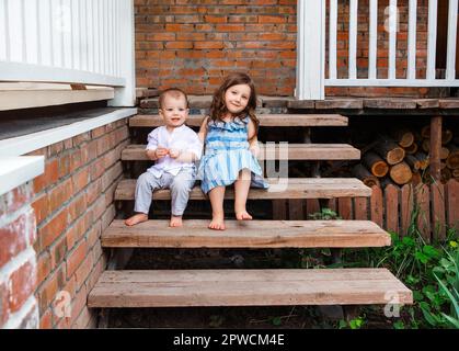 Mignon petit frère et soeur assis sur un escalier en bois près du porche de la maison de campagne et se détendre dans la zone rurale pendant les vacances d'été Banque D'Images