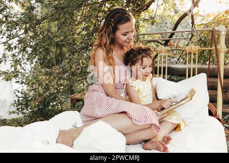 Belle mère et enfant passant du temps ensemble sur la terrasse ensoleillée près de la maison, maman lit des contes de fées à voix haute à la belle fille pendant que la petite fille s'assoit Banque D'Images