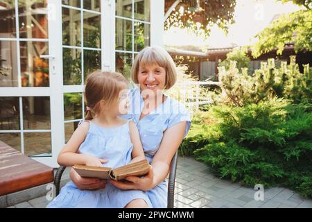 Grand-mère senior et petit-enfant passant du temps ensemble sur la terrasse ensoleillée près de la maison, grand-mère lit des contes de fées à haute voix à la belle fille tandis que petite fille Banque D'Images