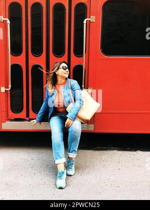 Bonne jeune femme voyageur en denim assis sur les marches du bus de voyage, regardant de côté avec le sourire tout en voyageant seule dans la ville d'Europe, en utilisant Banque D'Images