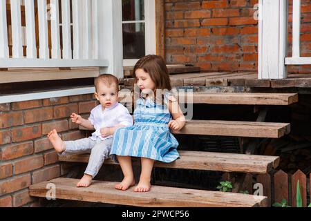 Mignon petit frère et soeur assis sur un escalier en bois près du porche de la maison de campagne et se détendre dans la zone rurale pendant les vacances d'été Banque D'Images