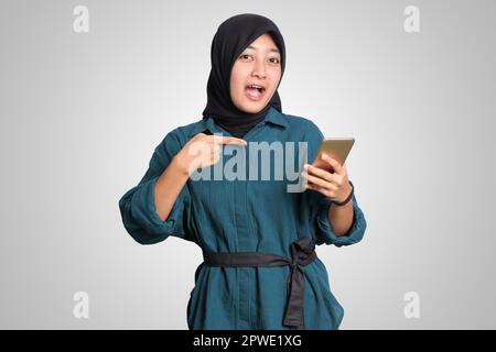 Portrait d'une femme musulmane asiatique excitée avec hijab tenant et touchant le téléphone mobile tout en pointant avec le doigt. Concept publicitaire. Image isolée activée Banque D'Images