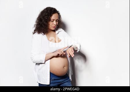 Portrait isolé sur fond blanc de la femme expectative aux cheveux bouclés comptant les contractions sur sa montre de poignet intelligente Banque D'Images