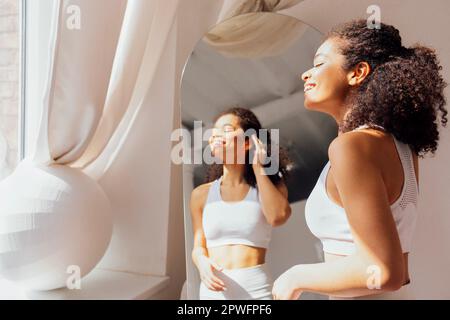 Jolie jeune femme de course mixte dans des vêtements blancs se tient près du miroir et des réflectes dedans. Mignonne souriante, cette jeune fille noire aux cheveux raillés profite de la lumière du soleil près du vent Banque D'Images