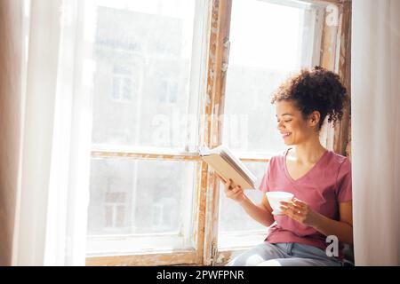 Jeune fille de race mixte souriante se trouve sur le rebord de la fenêtre avec une tasse de thé et de livre en porcelaine. Noir belle femme adolescente dans des vêtements décontractés lit un Banque D'Images