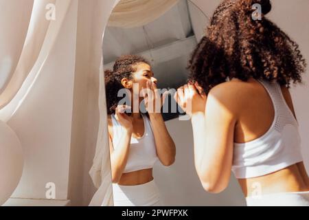 Jolie jeune femme de course mixte dans des vêtements blancs se tient près du miroir et des réflectes dedans. Mignonne souriante, cette jeune fille noire aux cheveux raillés profite de la lumière du soleil près du vent Banque D'Images