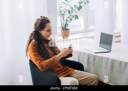 Travailleur à distance moderne : jeune femme heureuse utilisant un smartphone et un ordinateur portable pour l'apprentissage en ligne et le travail à distance à la maison. Jeune femme multitâche de Banque D'Images