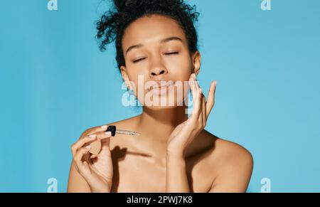 African American applique le sérum sur son visage avec une pipette, sur fond bleu. soins de la peau du visage avec traitement cosmétique huile et sérum Banque D'Images