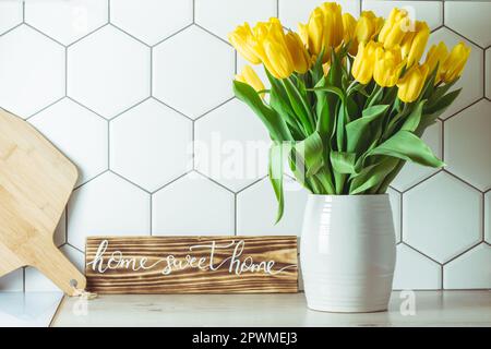 Vue de l'intérieur. Maison douce maison signe manuscrit sur le comptoir de cuisine à côté du bouquet de tulipe jaune dans le vase et tableau de coupe sur la tuile blanche hexagonale b Banque D'Images