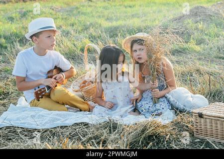 Portrait d'un garçon joyeux et de deux filles assis sur une couverture dans le champ, ayant pique-nique.Un moment de détente.Jouer de la guitare, ukulele, parler, plaisanter.Lumière ensoleillée Banque D'Images