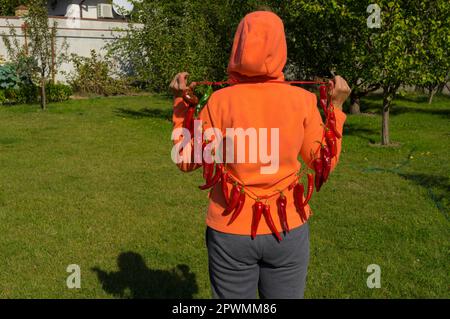 Vue arrière de moman ukrainien montrant un bouquet de piments rouges biologiques suspendus sur elle tout en se tenant dans le jardin Banque D'Images