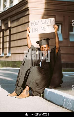 Un gars noir sans emploi assis avec une affiche en carton surélevé dans la rue à la recherche d'un emploi. Étudiant diplômé de l'université ou du collège en robe de troisième cycle et Banque D'Images