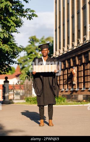 Jeune homme noir sans emploi debout avec une affiche en carton vide dans la rue à la recherche d'un emploi. Étudiant diplômé d'université ou d'université en robe de troisième cycle Banque D'Images