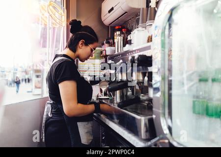 Le jeune barista féminin en gants prépare le café. Brunette girl verse du lait fouetté pour le cocktail dans une tasse en papier. L'intérieur du café de rue à emporter avec un prote Banque D'Images